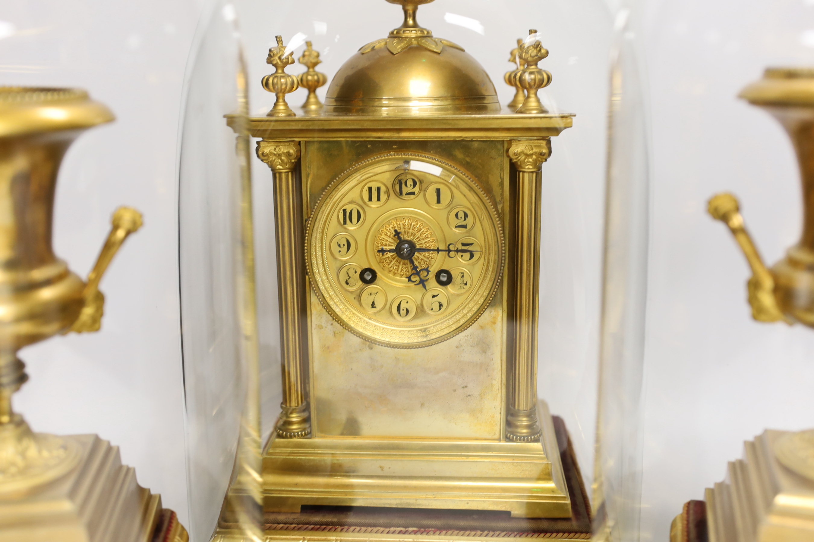 A gilt metal clock garniture, c.1900, with a pair of urns also under glass domes, French movement by A D Mougin, striking on a gong, (missing base for clock dome), 36cm high